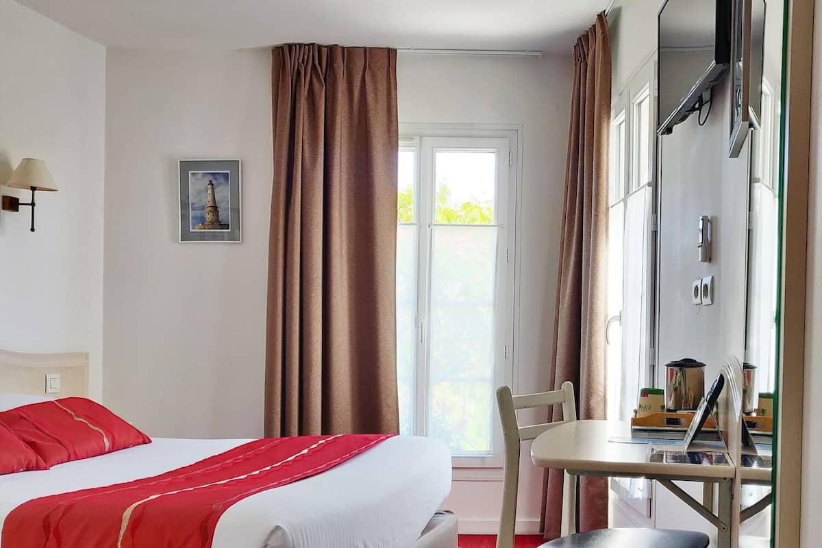 A hotel room with a bed, a desk, a chair, a trash can, and a window with curtains.