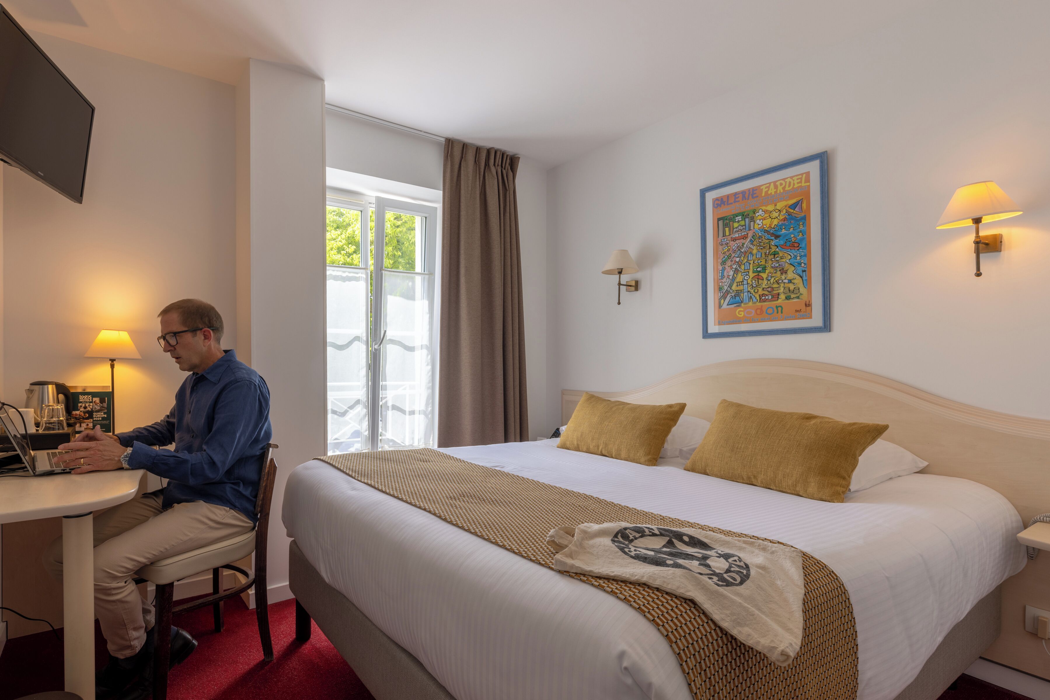 Ein Hotelzimmer mit einem Bett, einem Schreibtisch mit Laptop, einem Sessel, einem Fenster mit Vorhängen, einem an der Wand befestigten Fernseher und einem Wandbild.