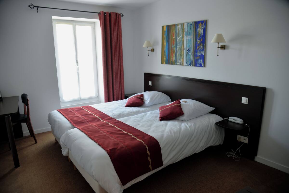 Quarto de hotel com uma cama grande, duas luminárias de cabeceira, mesa de cabeceira, escrivaninha e janela com cortinas.