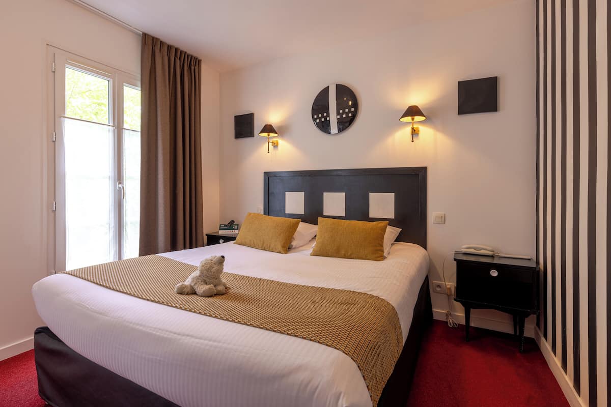 A hotel room with a large bed, two bedside lamps, a nightstand, and a teddy bear on the bed.