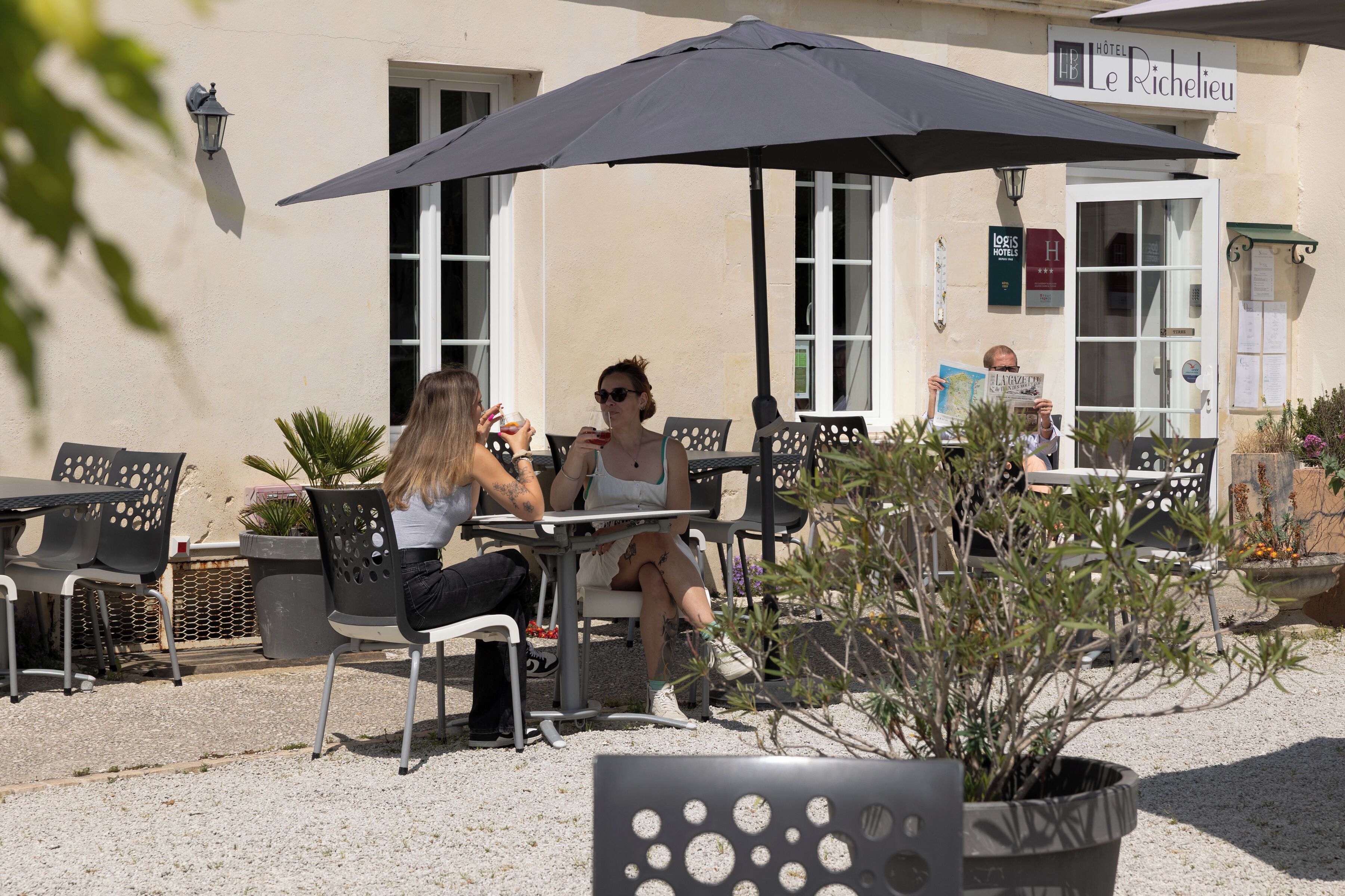 En udendørs café med borde og stole, en stor parasol og en bygning med skiltet "Hotel Le Richelieu".