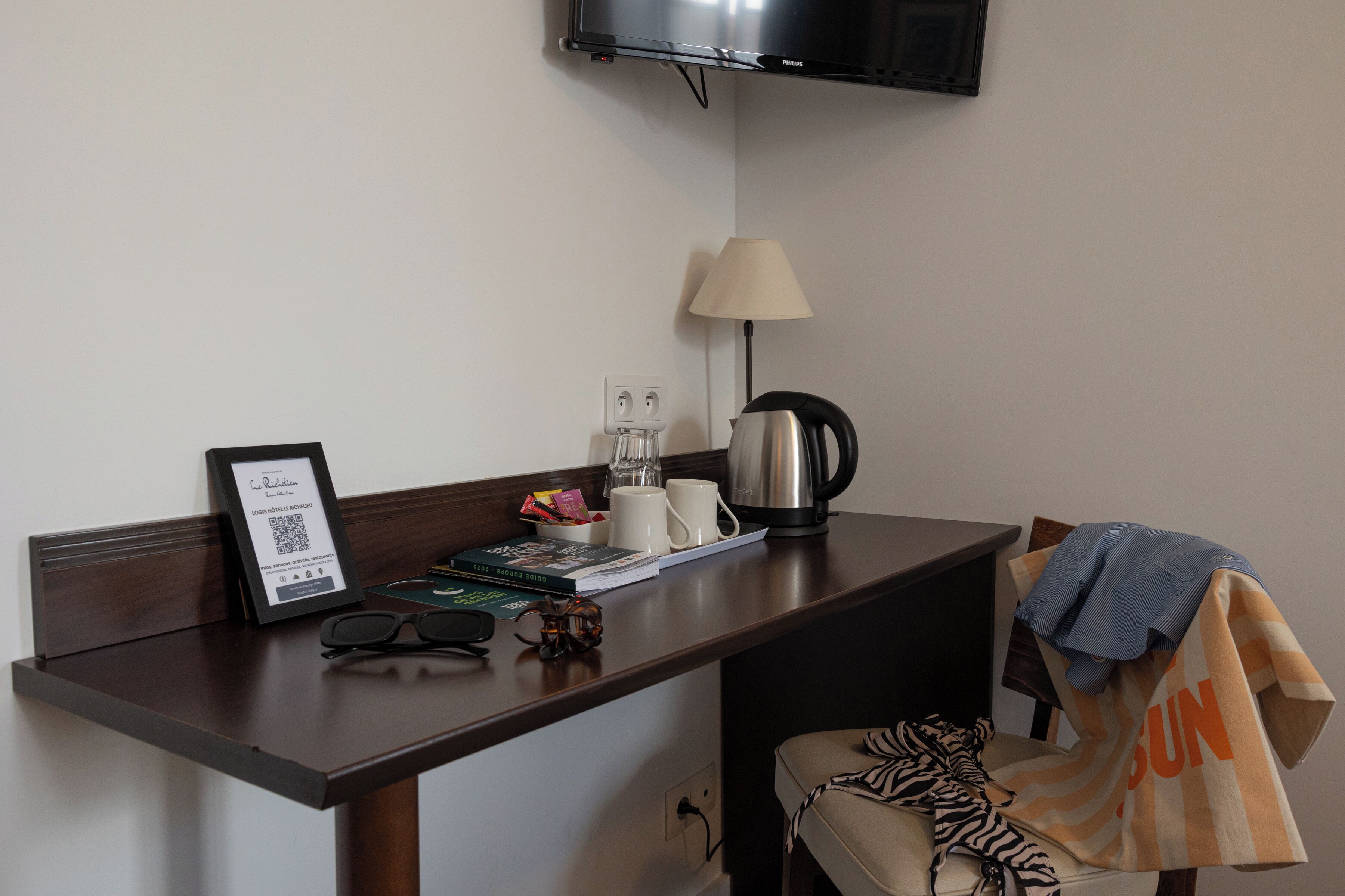 A hotel room with a desk, a lamp, a coffee maker, a framed certificate, a pair of glasses, and a chair with a blanket.