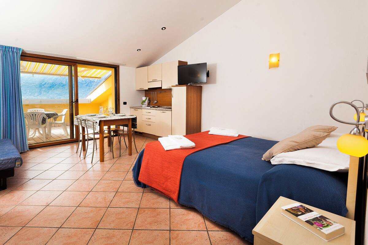 Une chambre avec un lit, un bureau et une chaise, un balcon avec une table et des chaises, et un coin cuisine.
