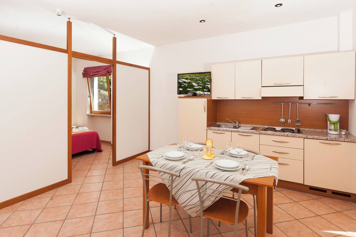 Une cuisine compacte avec un coin repas, dotée d’un sol carrelé, de meubles en bois et d’une fenêtre donnant sur un espace verdoyant.