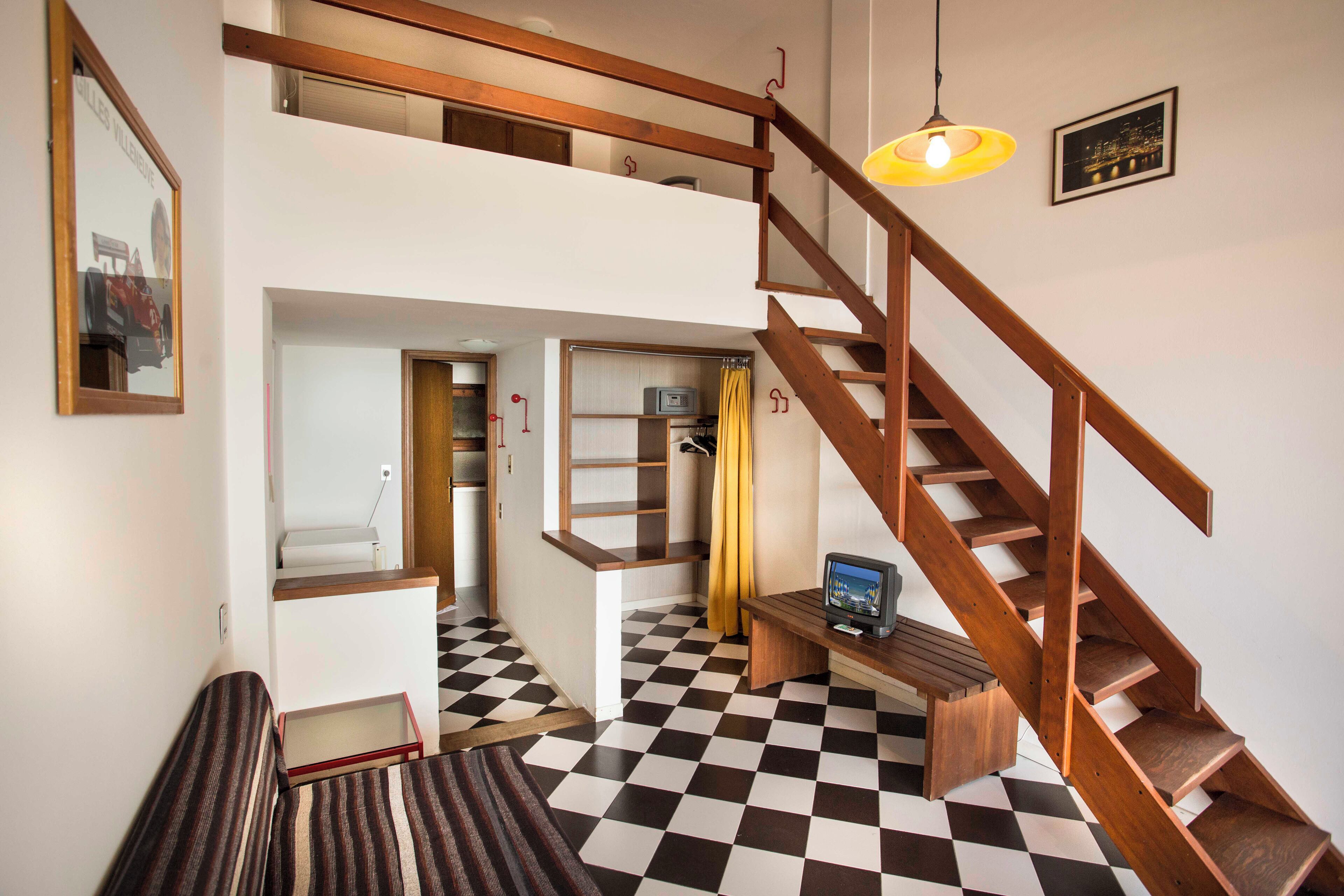 A room with a checkered floor, a wooden staircase, a television on a wooden stand, and a framed picture on the wall.