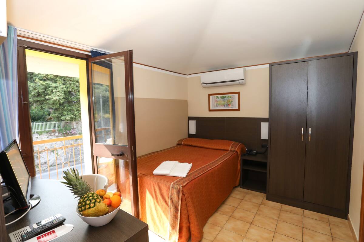 Une chambre d’hôtel avec un lit, un bureau équipé d’un ordinateur et de télécommandes, un panier de fruits et une armoire.