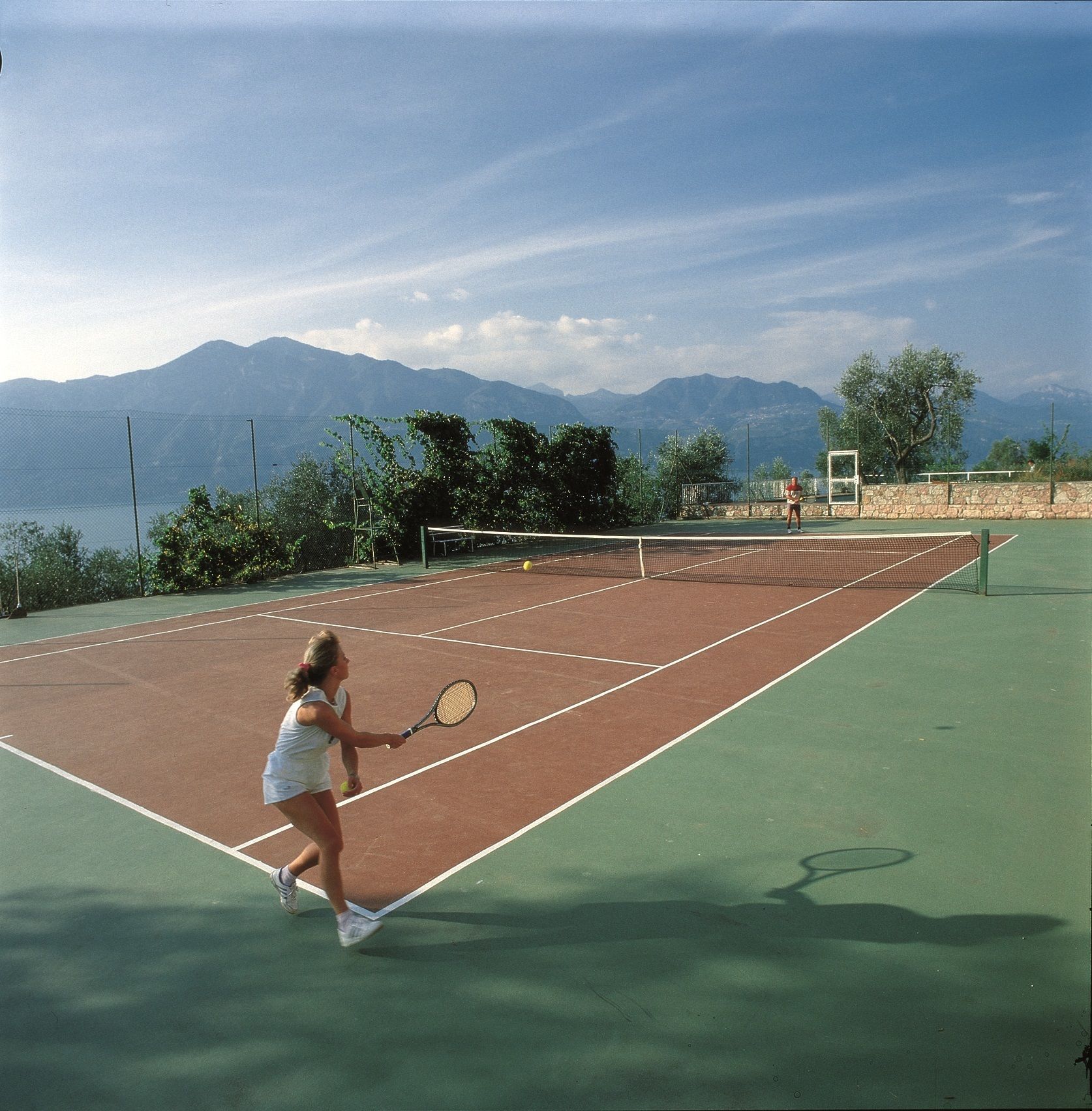 Ein Tennisplatz mit Netz, umgeben von einem Zaun und Bäumen.
