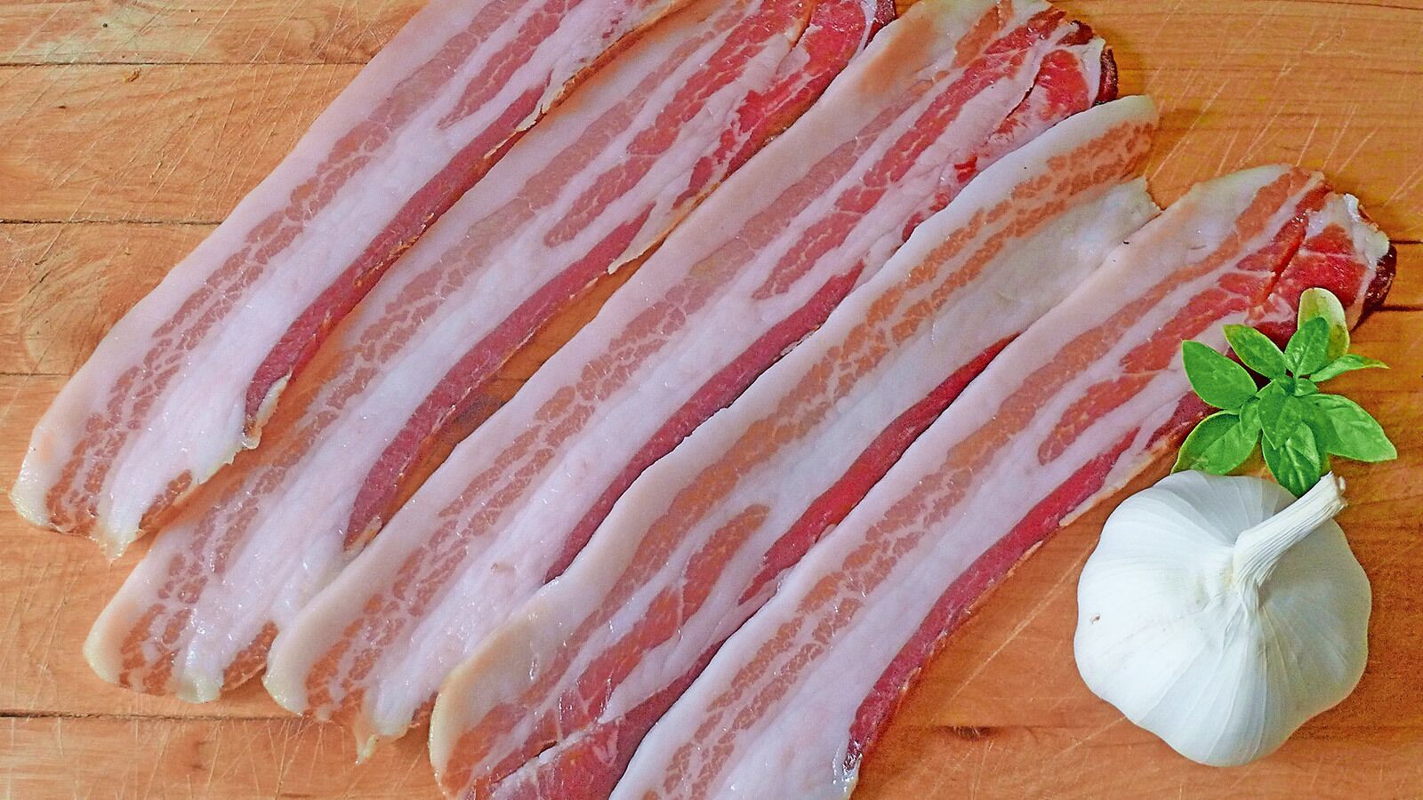 Raw bacon strips and a garlic bulb on a wooden surface.
