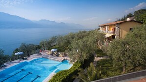 Una piscina con sillones reclinables y sombrillas, una casa con balcón con vistas a un lago, y montañas al fondo.