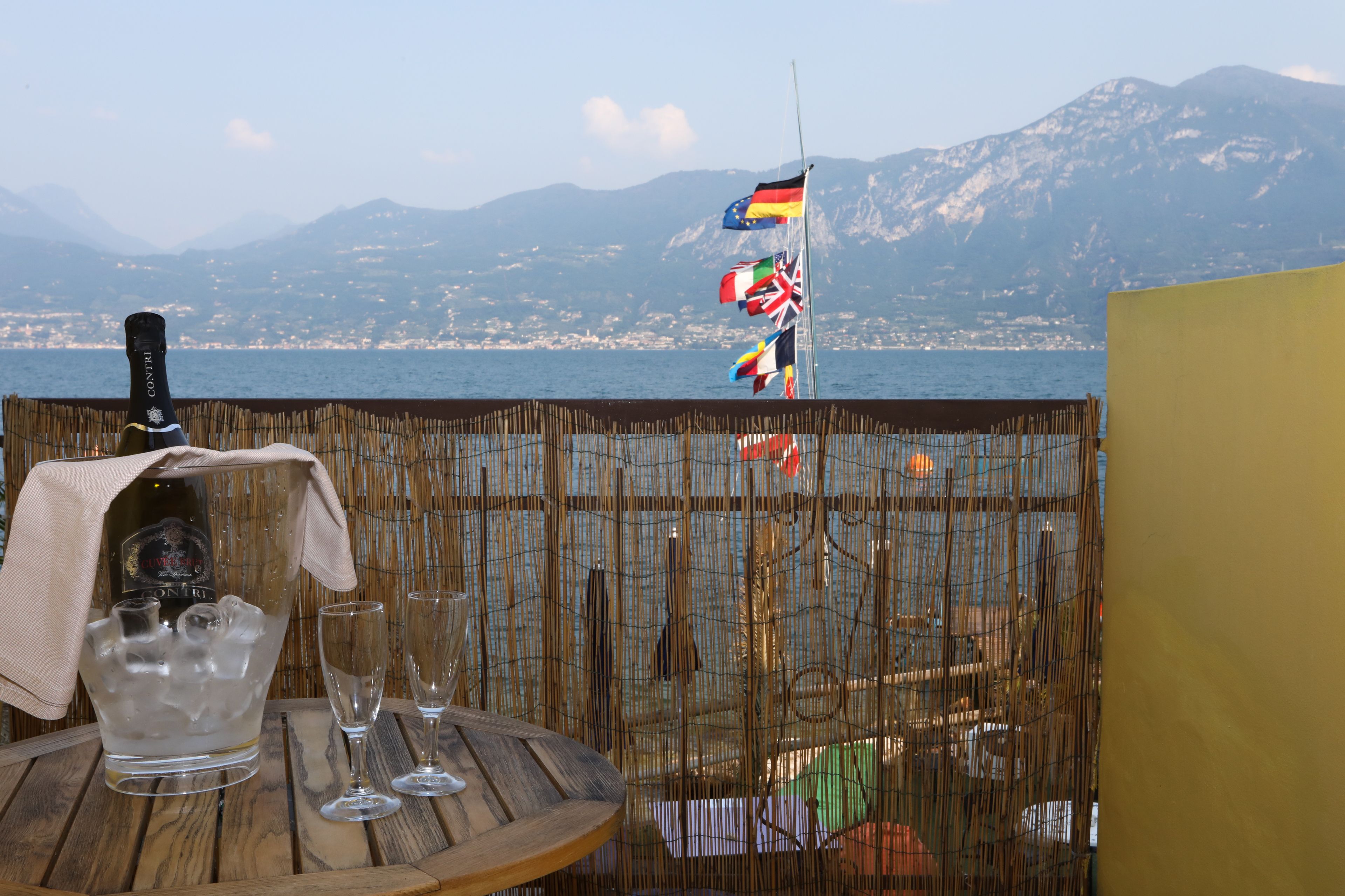 Ein Tisch mit einer Champagnerflasche, Eiswürfeln und Gläsern, mit Blick auf ein Gewässer, im Hintergrund Flaggen und Berge.