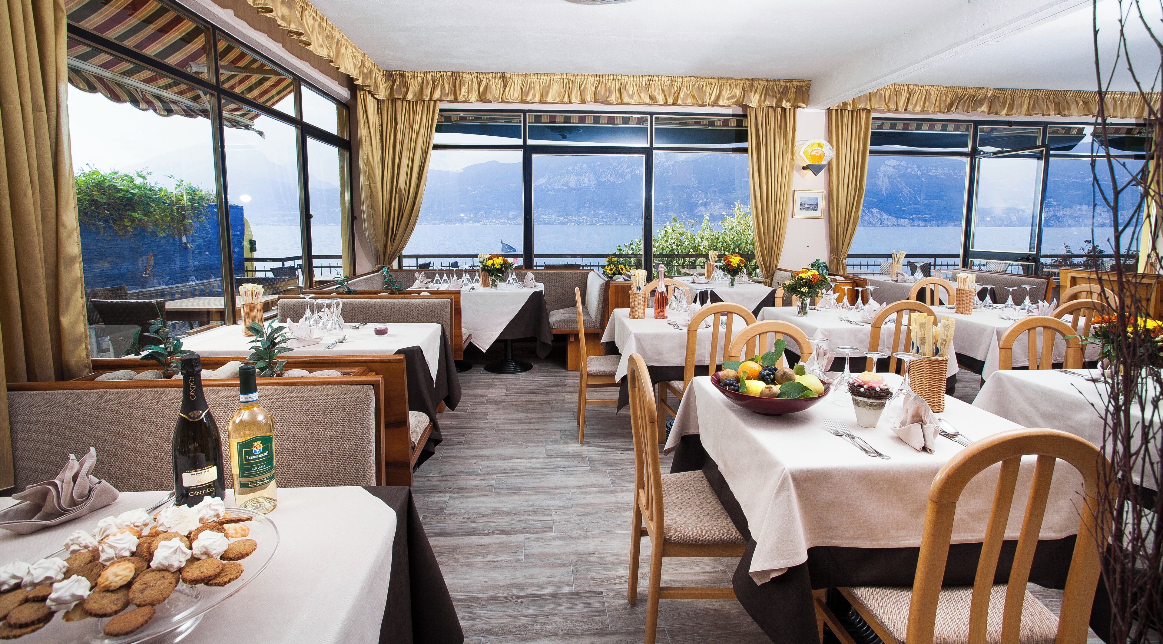 A restaurant with multiple tables, each set with white tablecloths, wooden chairs, and a variety of food and drink items.