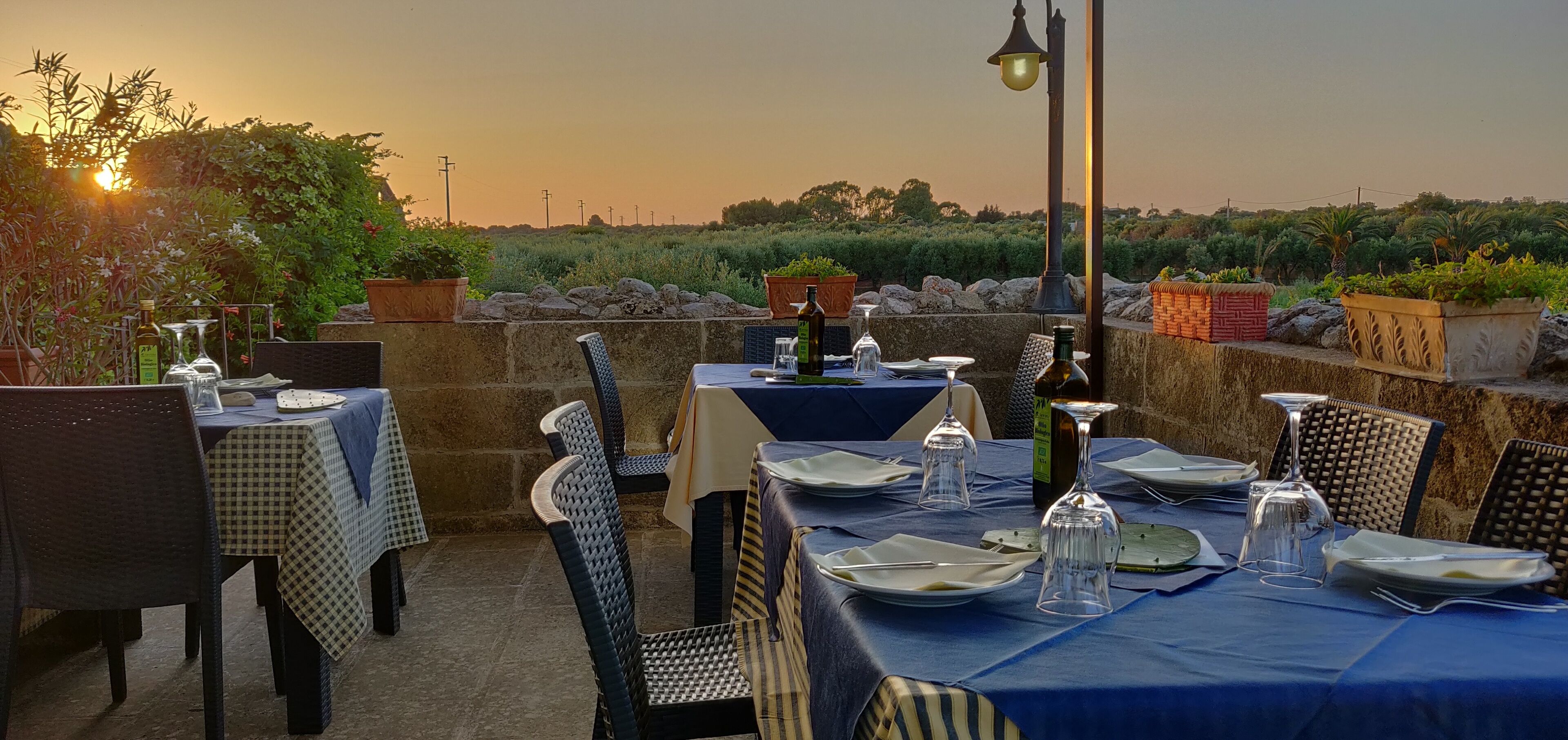 An outdoor dining area with tables set for two, surrounded by stone walls and potted plants.