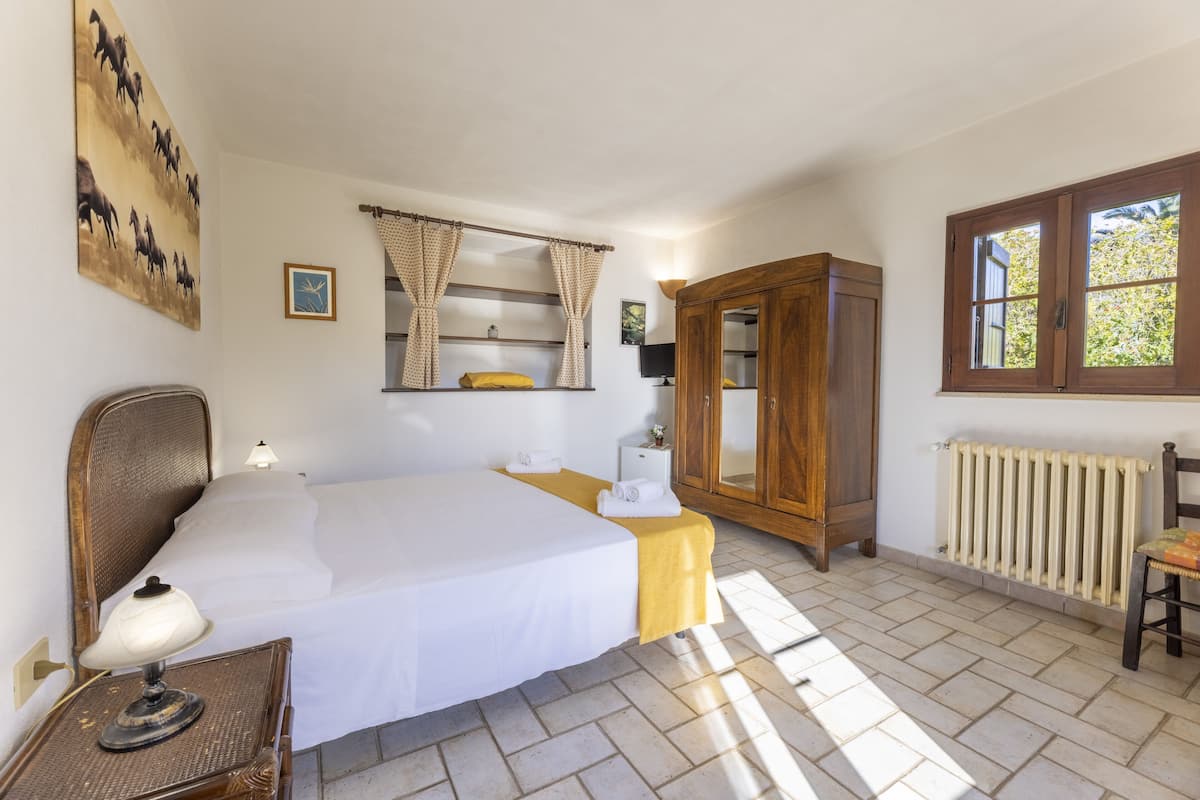 Une chambre à coucher comprenant un lit, une table de chevet, une armoire en bois, un radiateur et une fenêtre avec des rideaux.