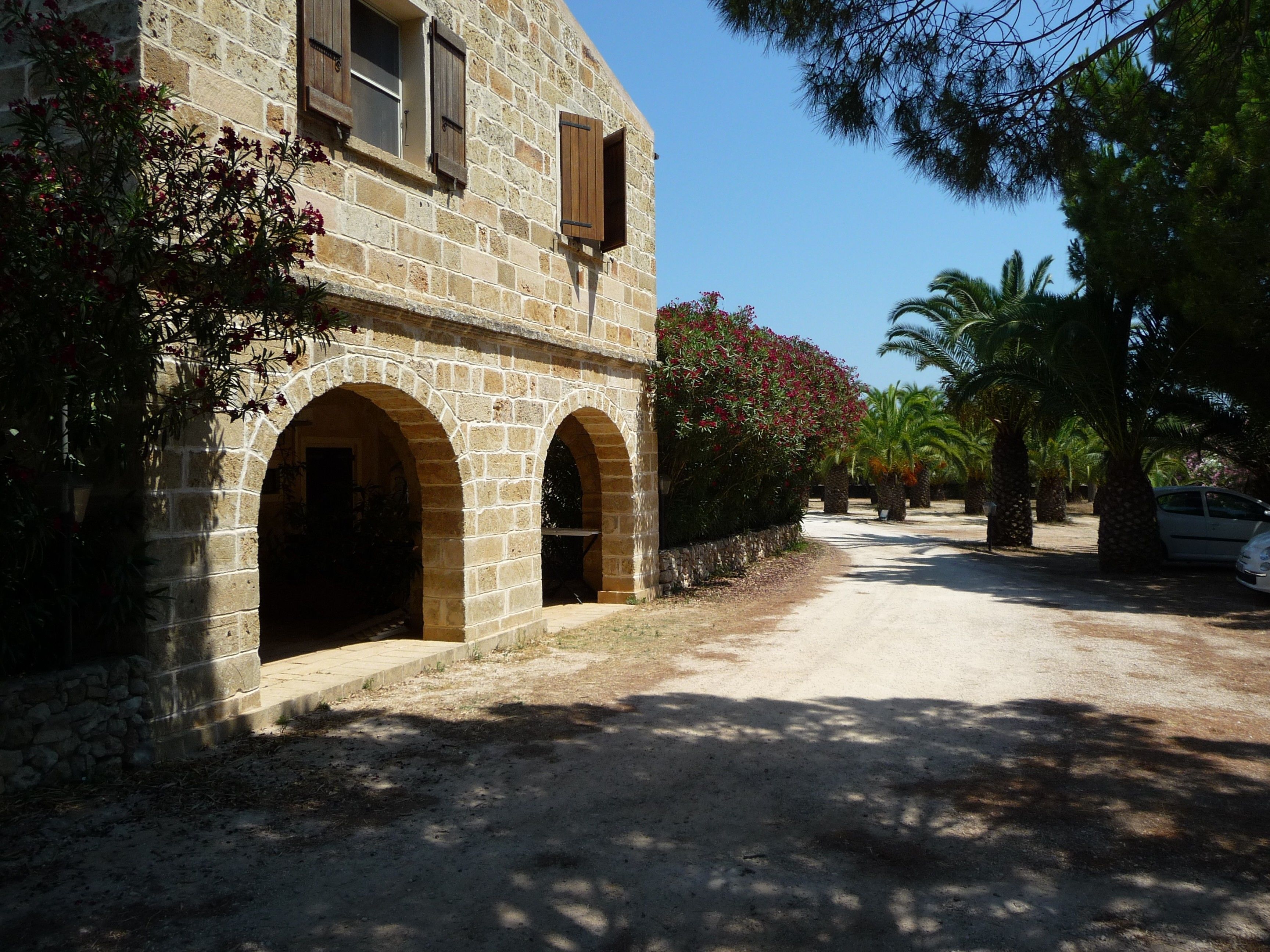 Un bâtiment en pierre, doté d’arcades au niveau des portes et des fenêtres, entouré de palmiers et bordé d’un chemin de terre.