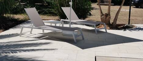 Zona junto a la piscina con sillas de descanso blancas y un gran paraguas, rodeada de vegetación y un edificio al fondo.