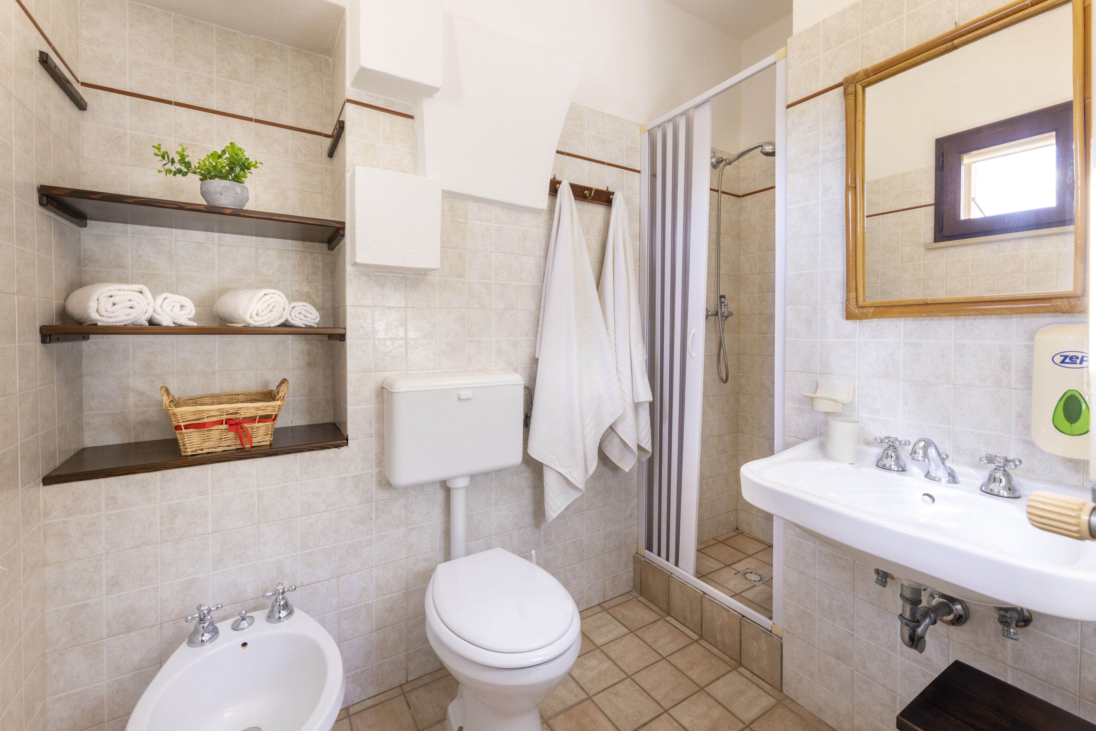 Un baño con inodoro, bidé, lavamanos, repisas con toallas y una cesta, zona de ducha y un espejo.