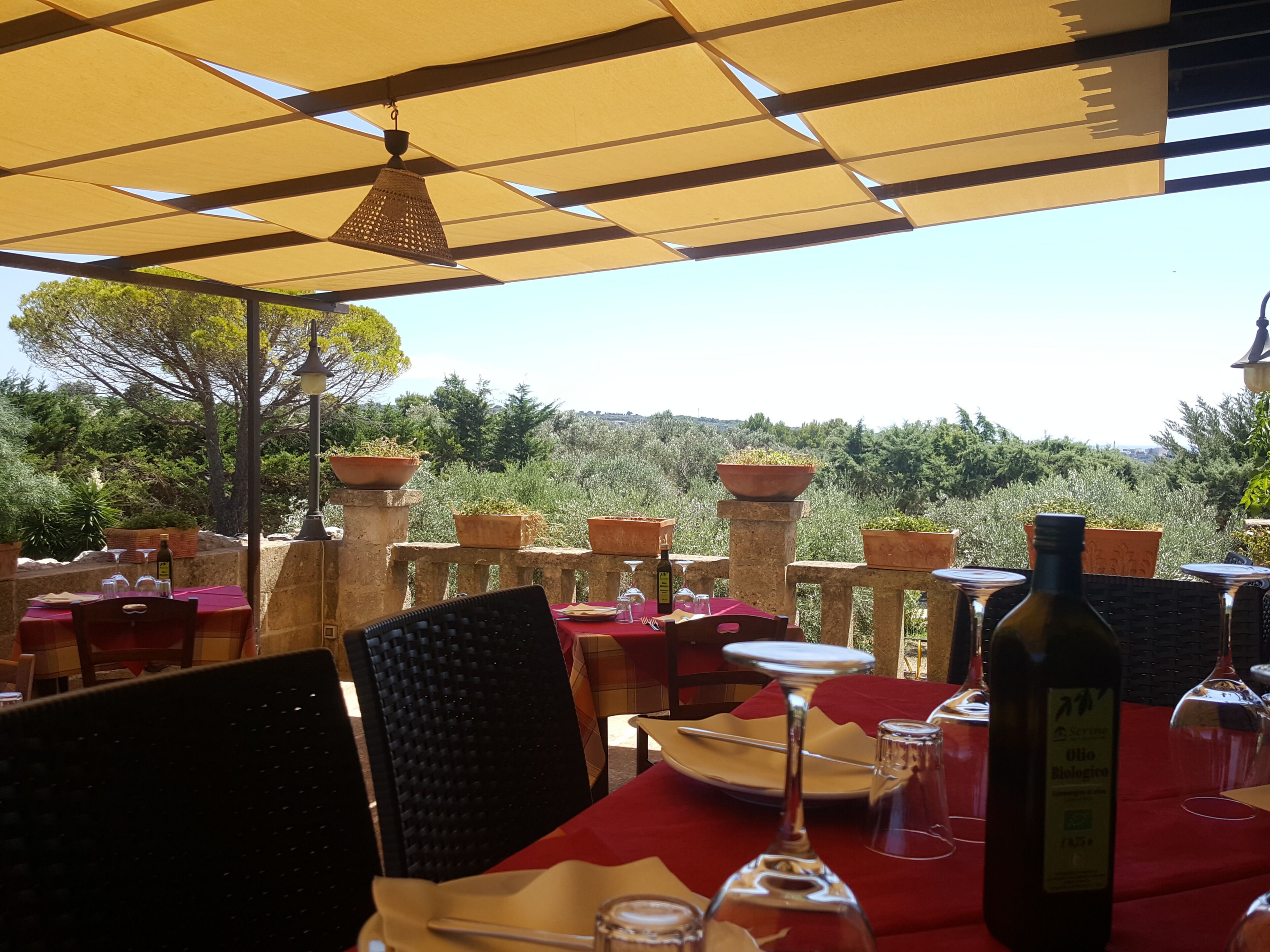 Un área de comedor al aire libre con mesas dispuestas para dos, con mantel rojo, copas de vino y una botella de vino.
