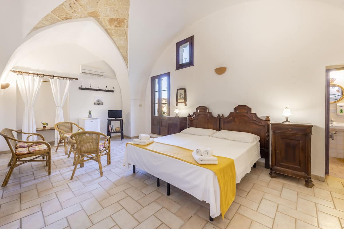Une chambre d’hôtel avec un lit, deux fauteuils en osier, une table de chevet en bois et une petite table avec une lampe.