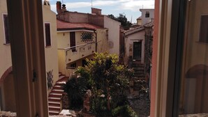 A view through a window of a courtyard with a staircase leading to a building, surrounded by greenery and other structures. - B&B Casa Saba (Orosei)