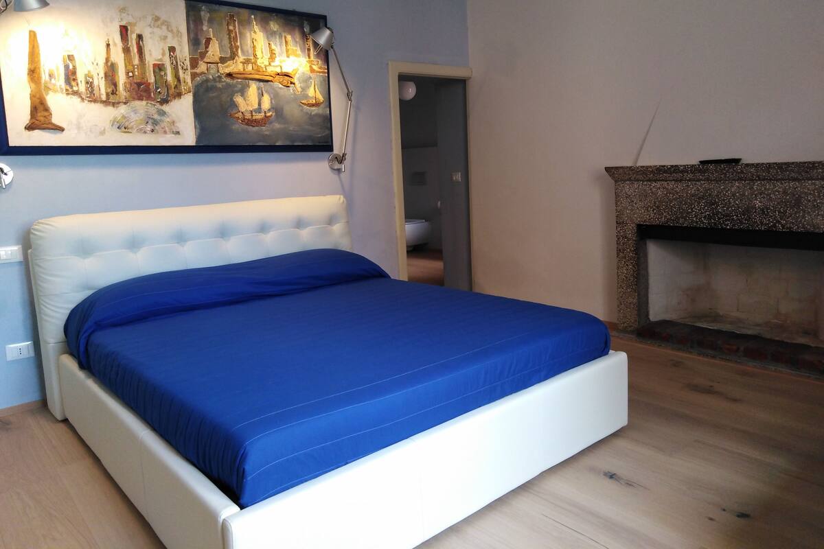 A bedroom with a white bed, blue bedding, a painting above the bed, a lamp, and a fireplace.