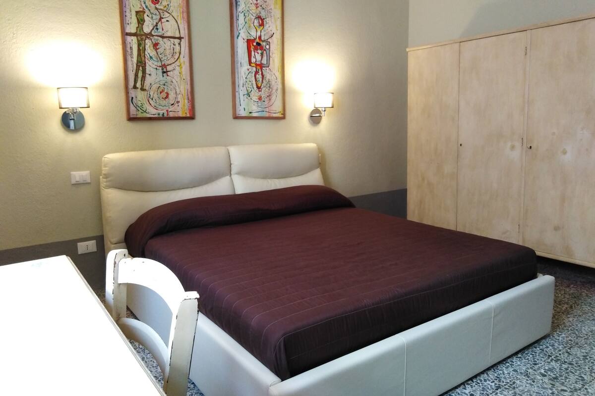A bedroom with a bed, a wardrobe, two wall-mounted lamps, and two abstract paintings.