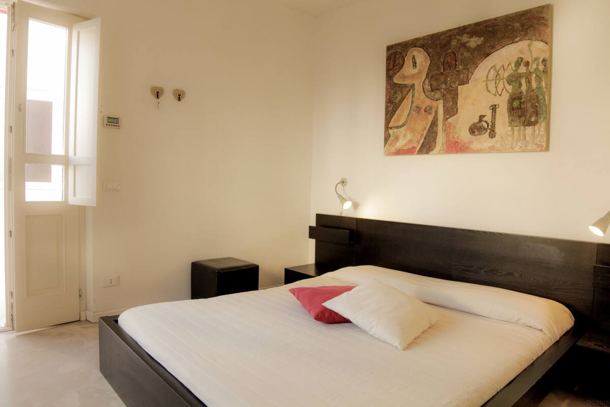 A bedroom with a large bed, a nightstand, a wall-mounted lamp, and a door.