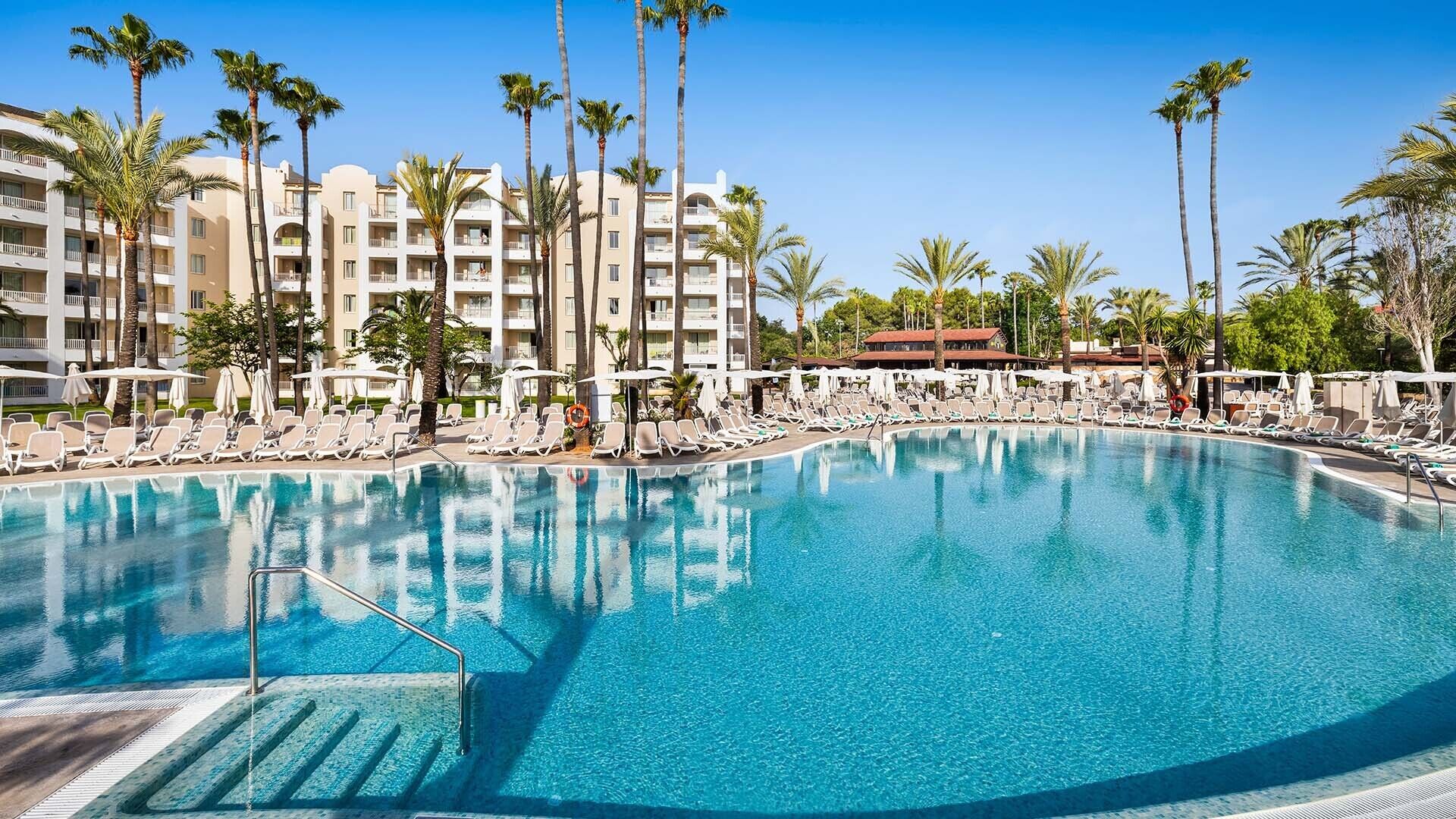 Ein großer, klarer Pool, umgeben von Liegestühlen und Palmen, mit einem mehrstöckigen Hotel im Hintergrund.
