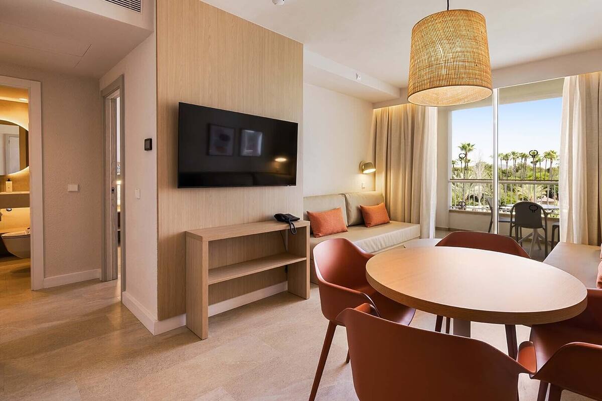 A modern hotel room with a flat-screen TV, a round dining table with chairs, a sofa, and a large window with curtains.