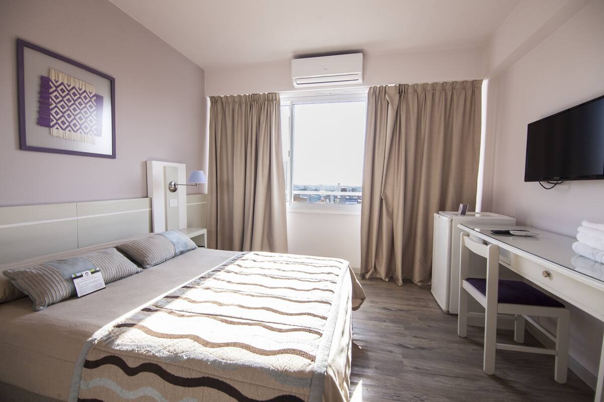 Hotellihuone, jossa on sänky, työpöytä, tuoli, televisio ja ikkuna, jossa on verhot.