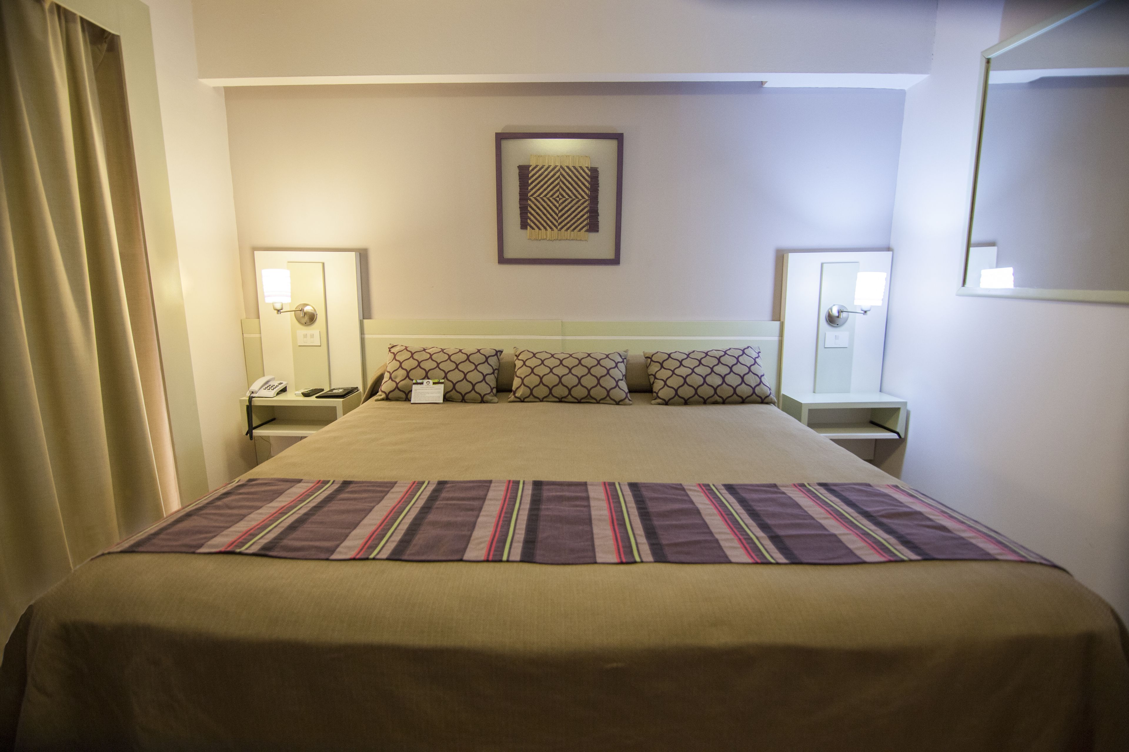 Ein Hotelzimmer mit einem großen Bett, einem Nachttisch, einem Spiegel an der Wand und einem gerahmten Kunstwerk über dem Bett.