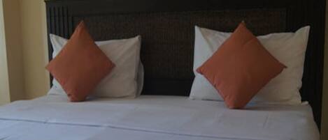 Una cama bien hecha con ropa blanca y dos almohadas naranjas, una toalla doblada y dos cuadros en la pared.