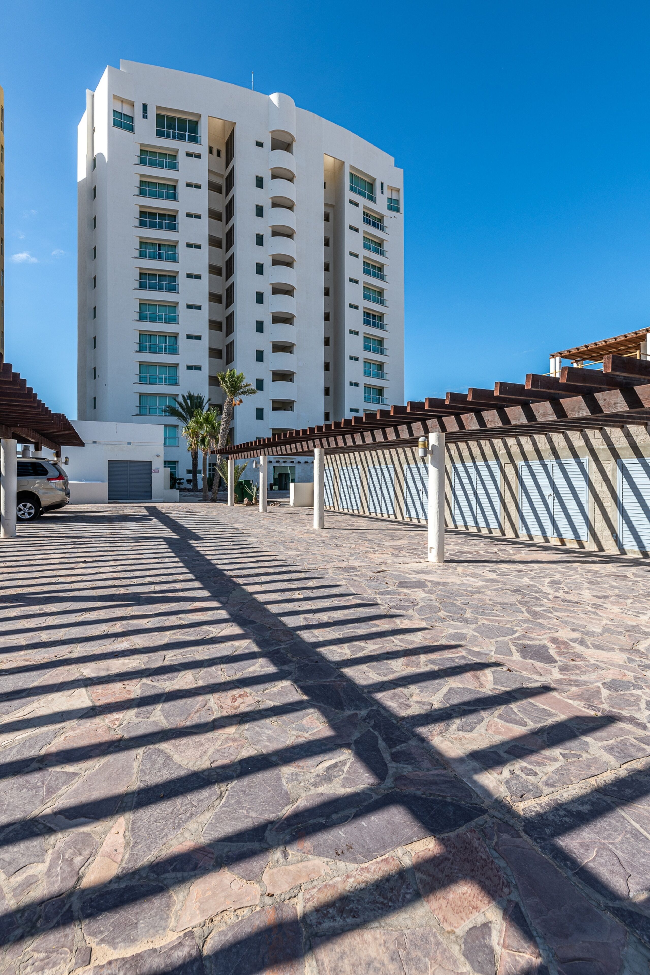 A stone-paved area with a car parked under a pergola, a tall white building with multiple balconies, and a clear blue sky.