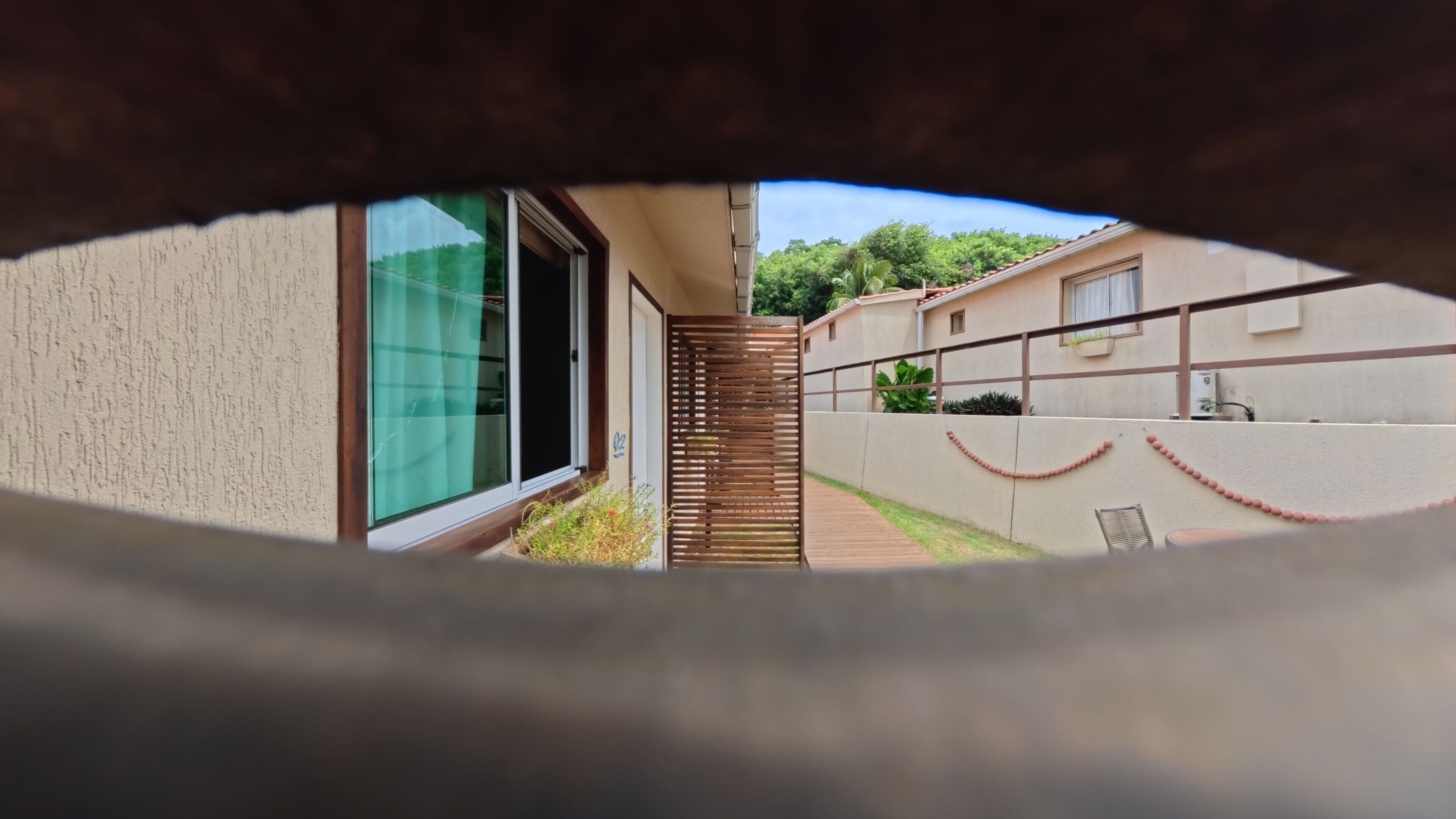 Visão pela janela de um edifício, com porta de vidro, cerca de madeira e caminho que leva a um edifício com fachada branca.