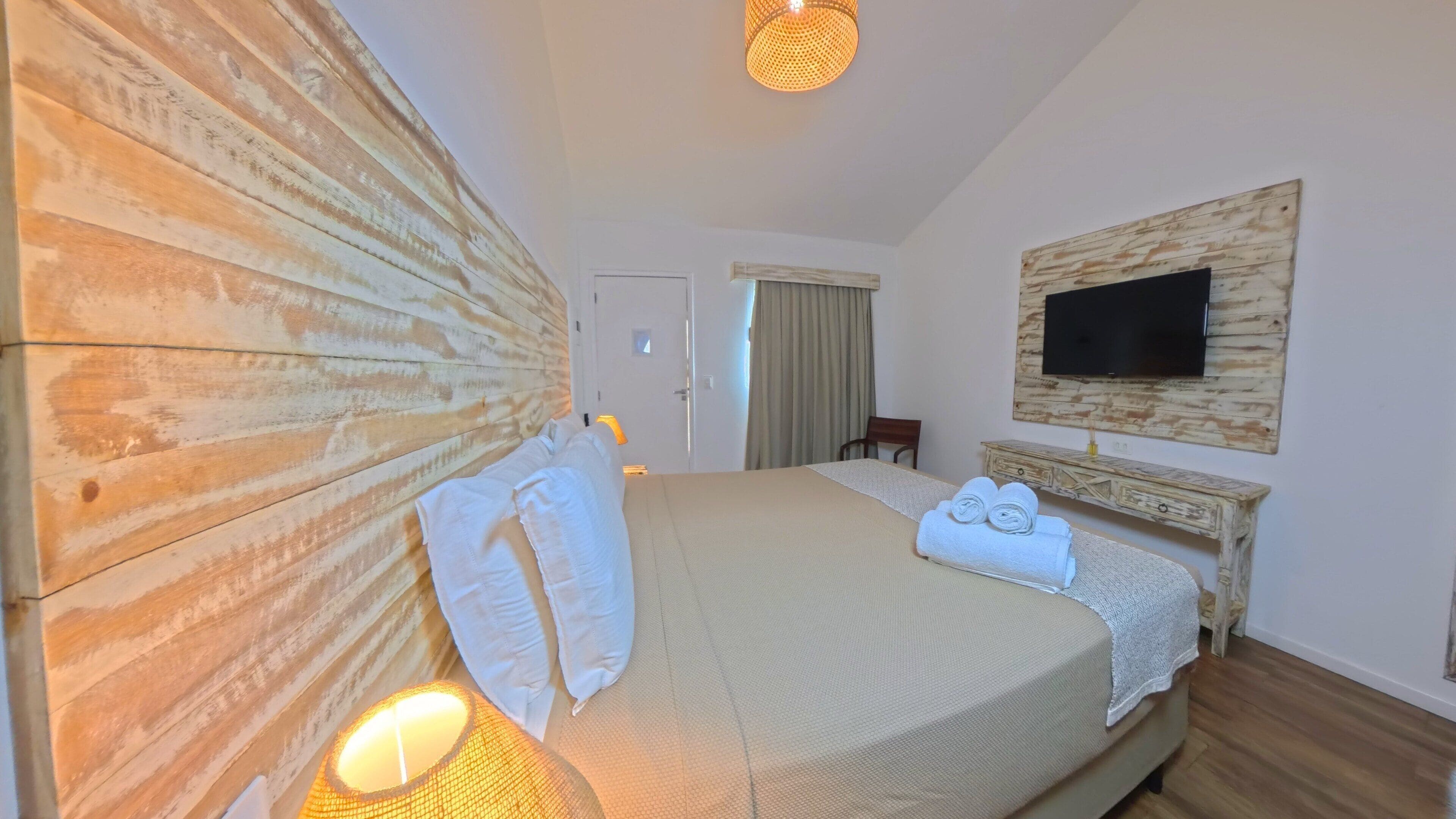 Ein Hotelzimmer mit einem großen Bett, einem an der Wand befestigten Fernseher, einem hölzernen Kopfteil und einem Nachttisch mit einer Lampe.