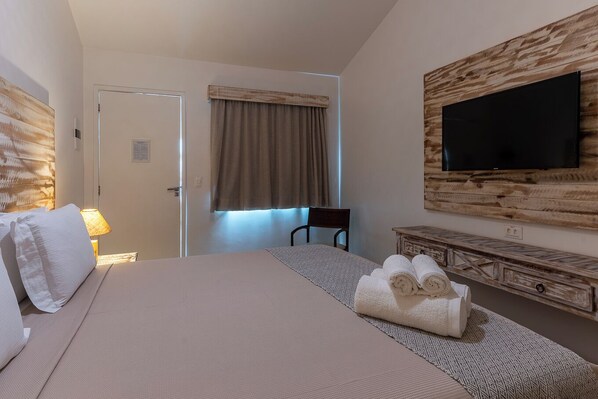 A hotel room with a large bed, a flat-screen TV mounted on a wooden wall, a desk with a chair, and a window with curtains. - Pousada Tubarão (Fernando de Noronha)