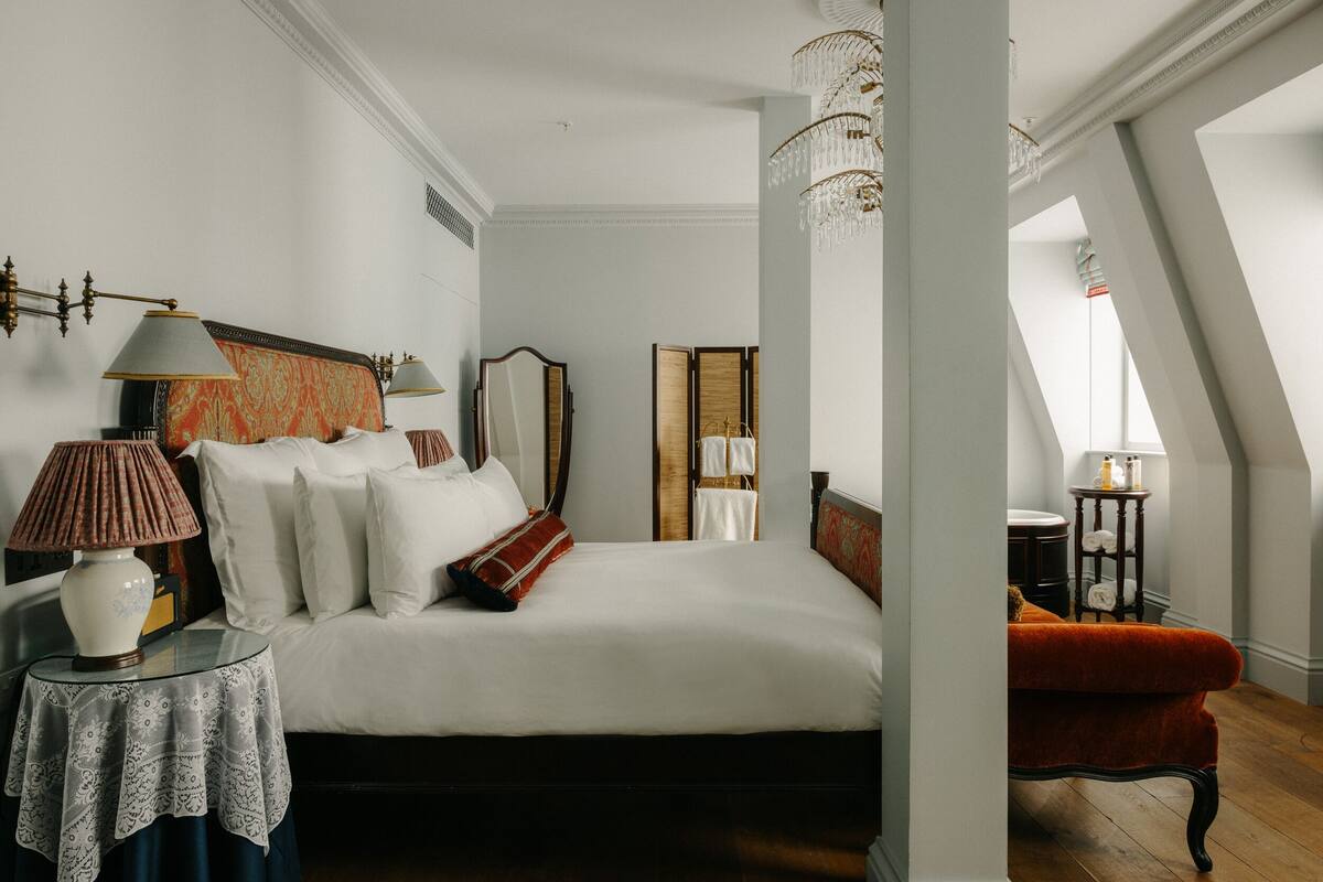 Een slaapkamer met een groot bed, een nachtkastje met een lamp, een spiegel en een rode stoel.