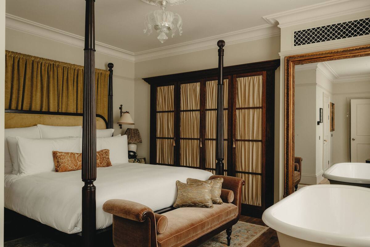Een slaapkamer met een hemelbed, een zithoek met een bank, een groot raam met gordijnen en een bad.
