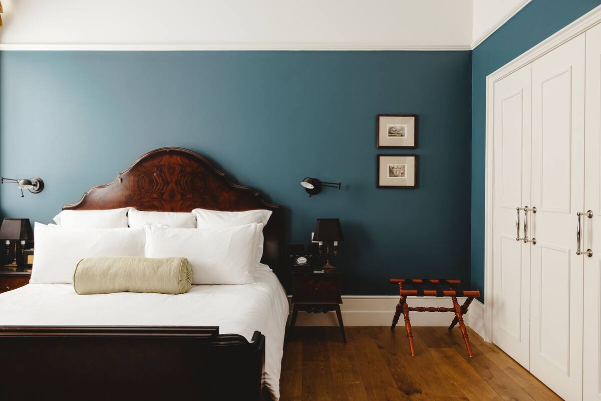 Een opgemaakt bed met witte lakens en een beige sierkussen in een kamer met donkerblauwe wanden en houten vloeren.
