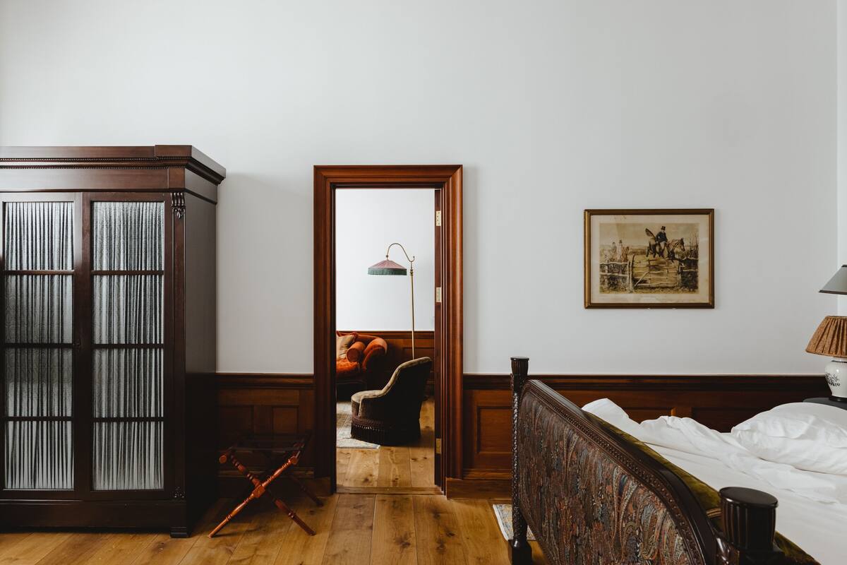 Een kamer met een houten garderobe, een bed met hoofdeinde, een stoel, een schilderij aan de muur en een lamp op een nachtkastje.