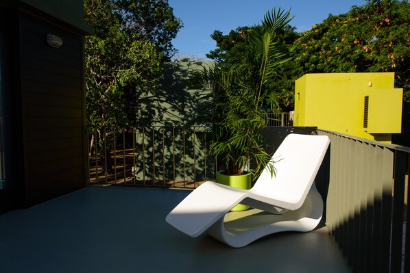 Un espace de détente extérieur moderne, avec un fauteuil blanc, un pot de fleurs vert contenant un palmier, et un bâtiment jaune en arrière-plan.