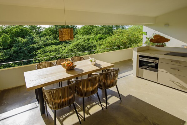 Un balcón con una mesa de comedor, sillas de mimbre y vistas a abundante vegetación.