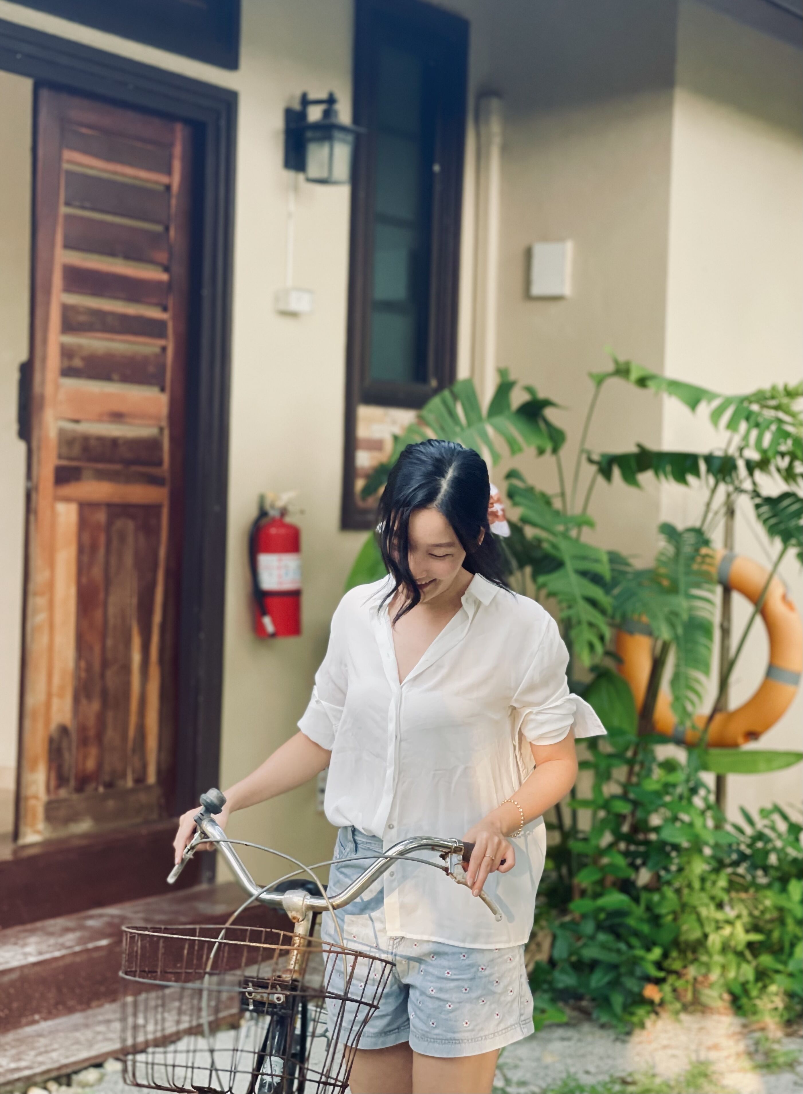 A person standing next to a bicycle with a basket, in front of a building with a wooden door and a fire extinguisher mounted on the wall.