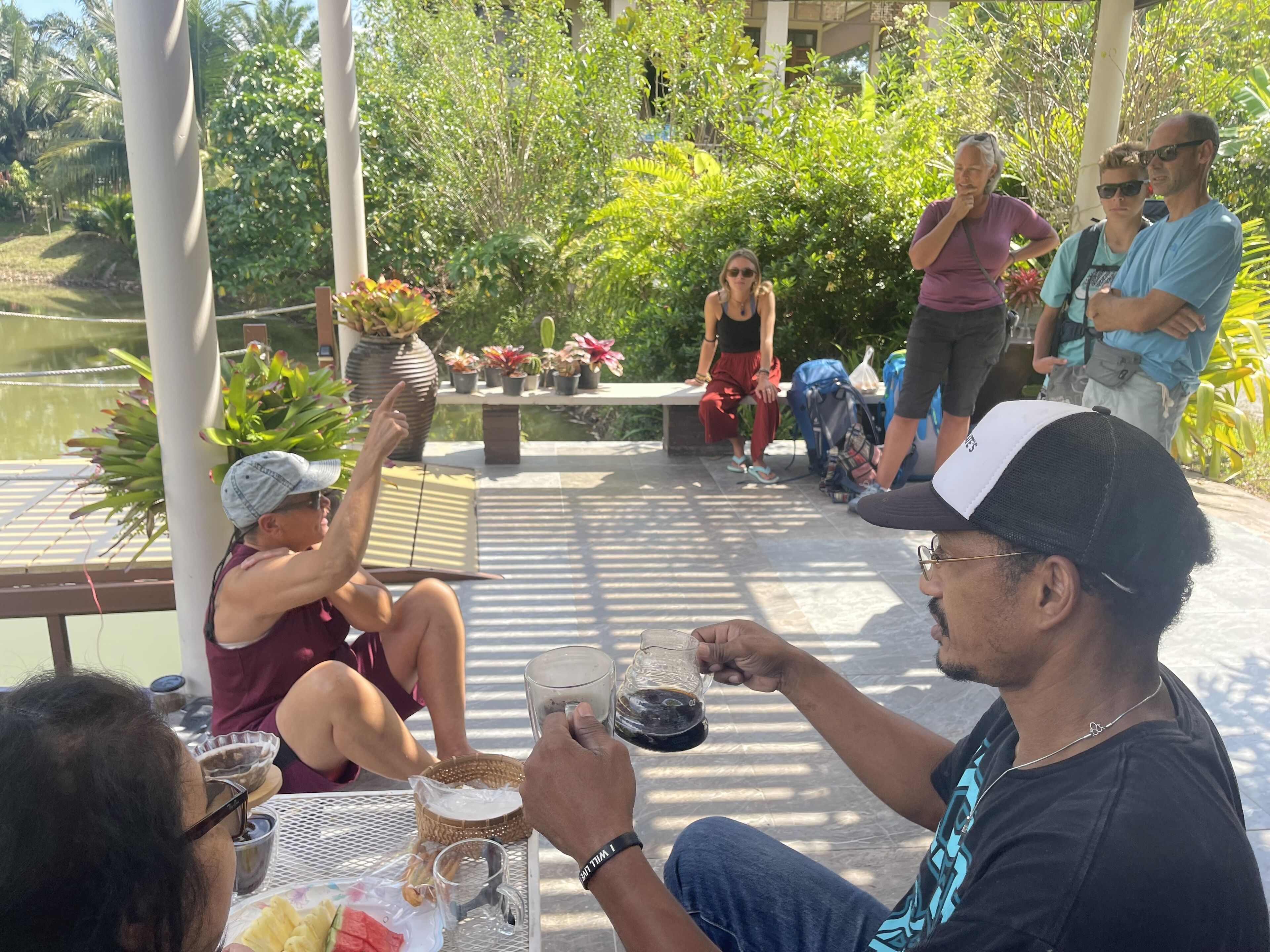 A group of people sitting at an outdoor table with drinks, surrounded by greenery and a body of water.