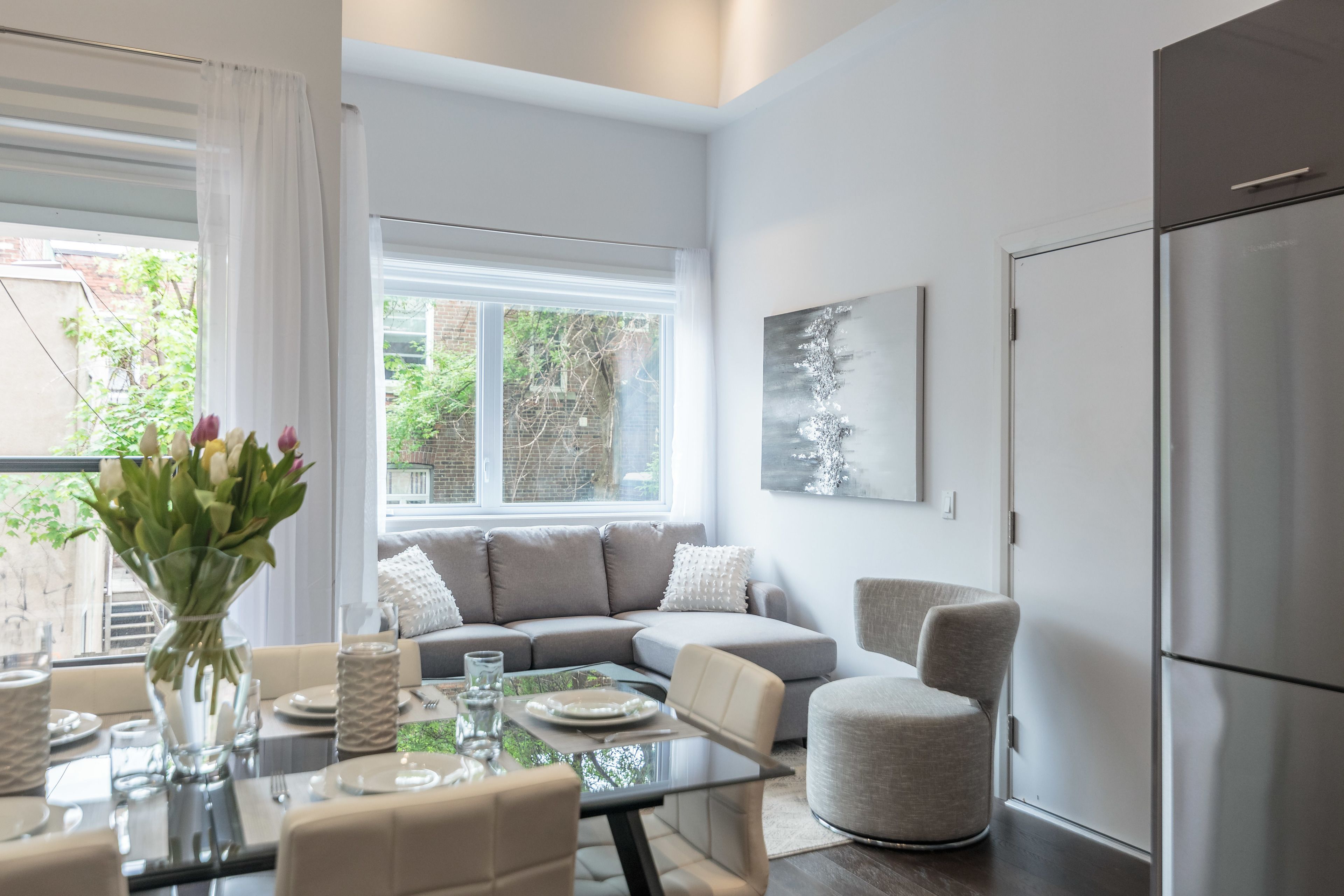 Una sala de estar moderna con una mesa de comedor de vidrio, sofás grises y un ventanal con luz natural.