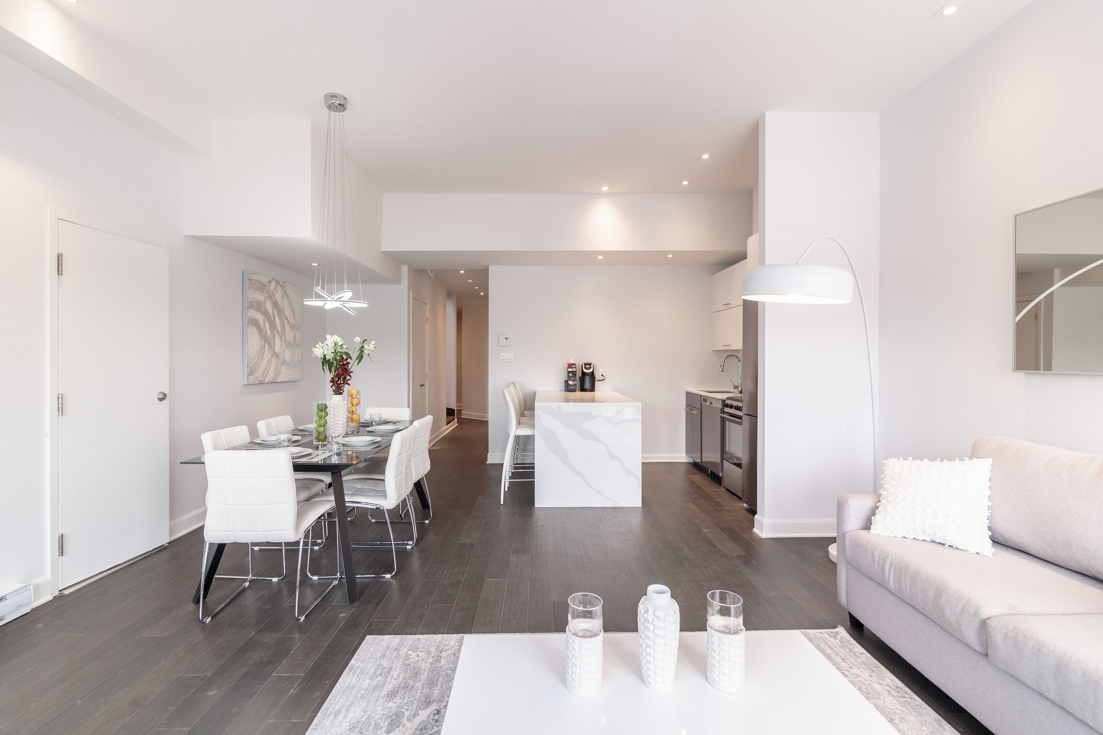 A modern living room with a white sofa, a dining area with white chairs, a kitchen with white cabinets, and a hallway with a white door.
