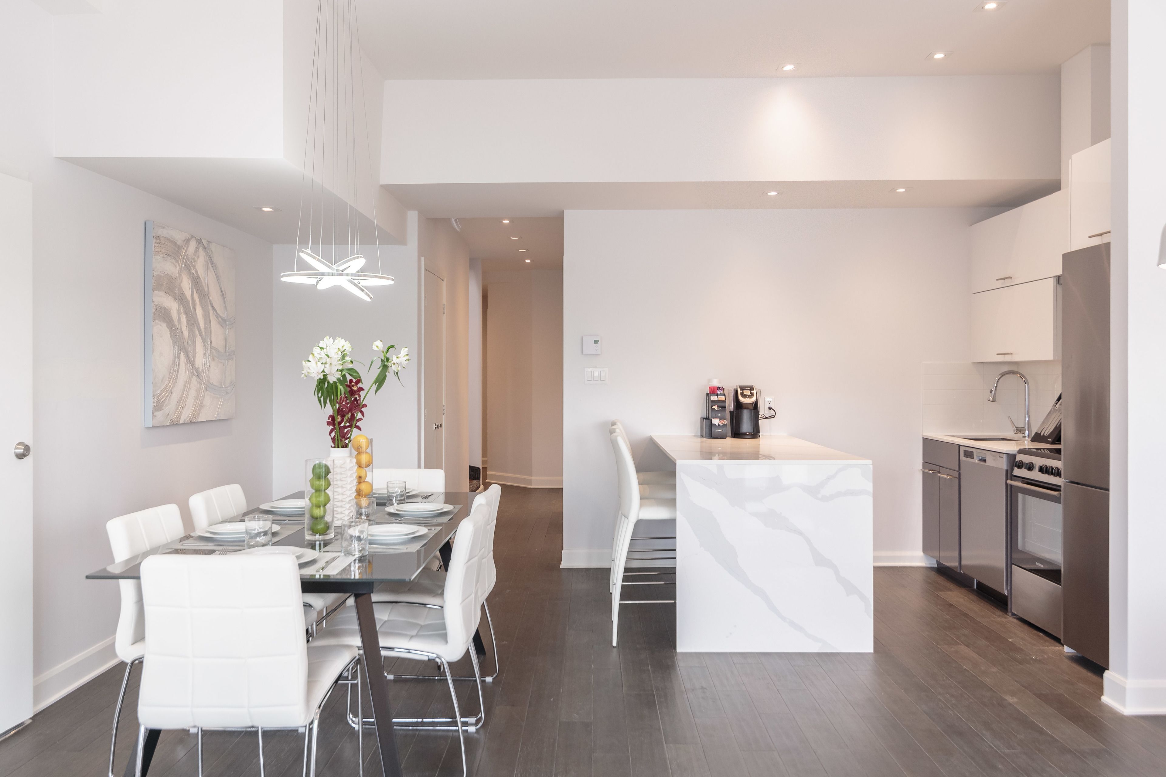 Una cocina moderna con electrodomésticos de acero inoxidable, un comedor con sillas blancas y una mesa de vidrio, y un pasillo que lleva a un dormitorio.