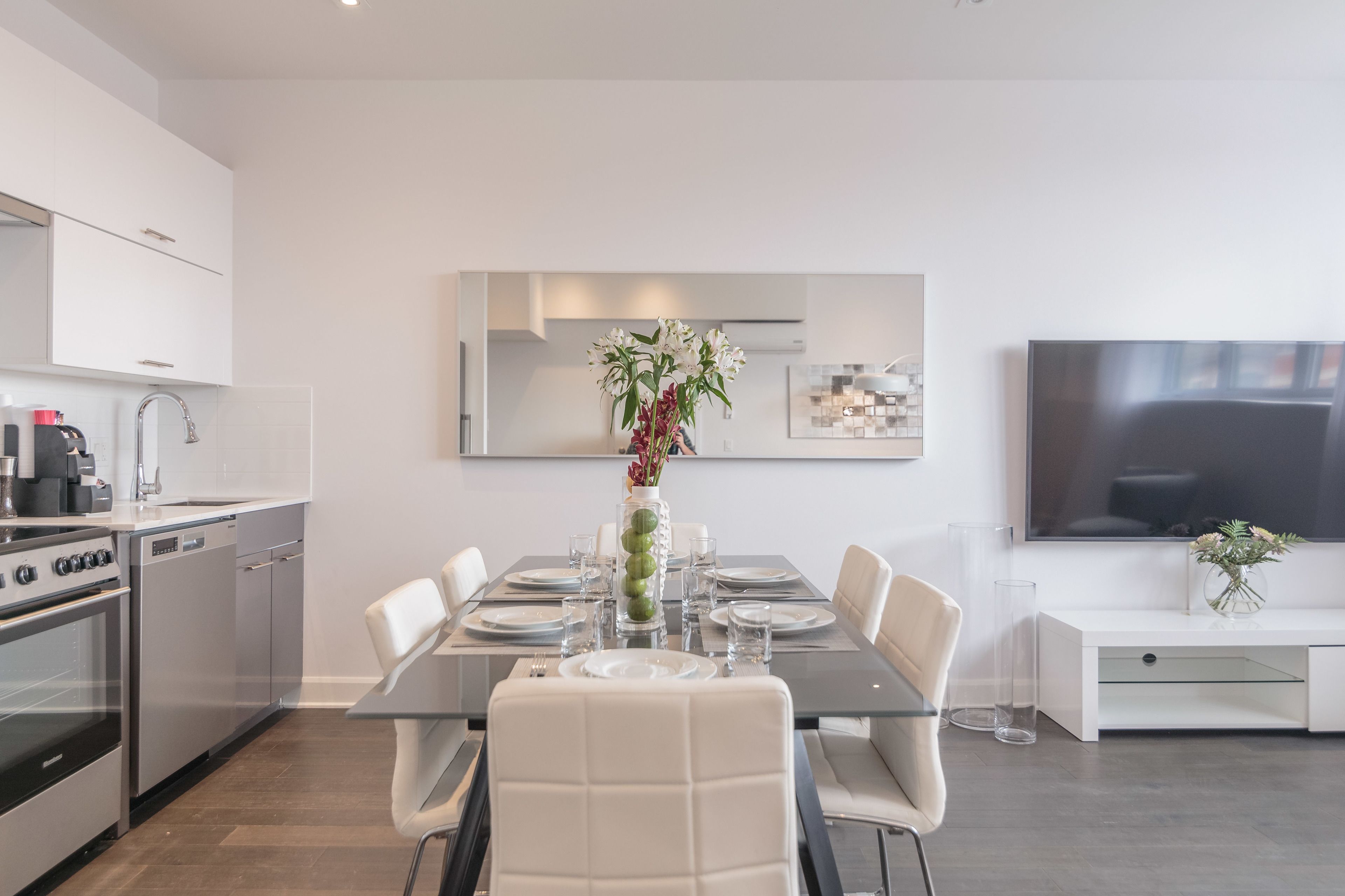 Una cocina moderna con zona de comedor, muebles blancos, electrodomésticos de acero inoxidable y un televisor de pantalla plana.