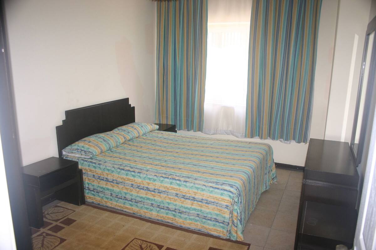 A bedroom with a bed, a nightstand, a dresser, and a window with curtains.