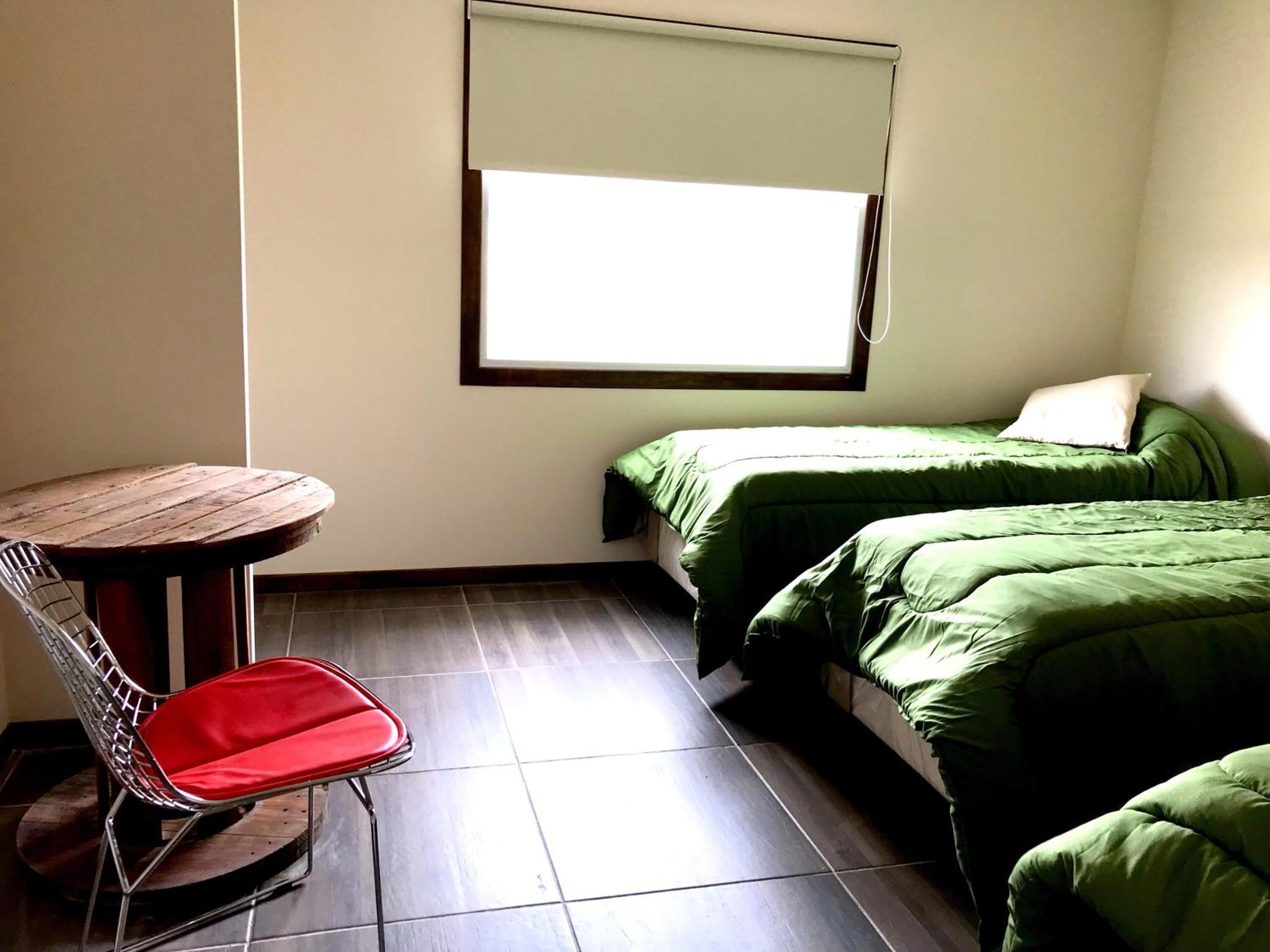Una habitación pequeña y funcional con una mesa redonda de madera, una silla roja, dos camas individuales con ropa de cama verde y un ventanal con persiana.