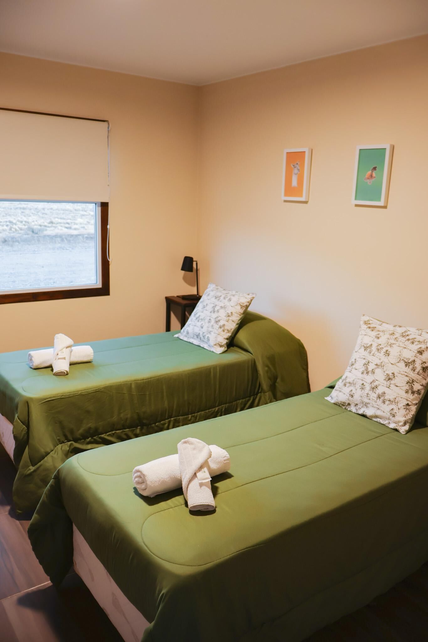 Een kamer met twee bedden, elk met een groene spreidek en een kussen. Er is een raam met een blind, een kleine tafel met een lamp, en twee ingelijste foto’s aan de muur.